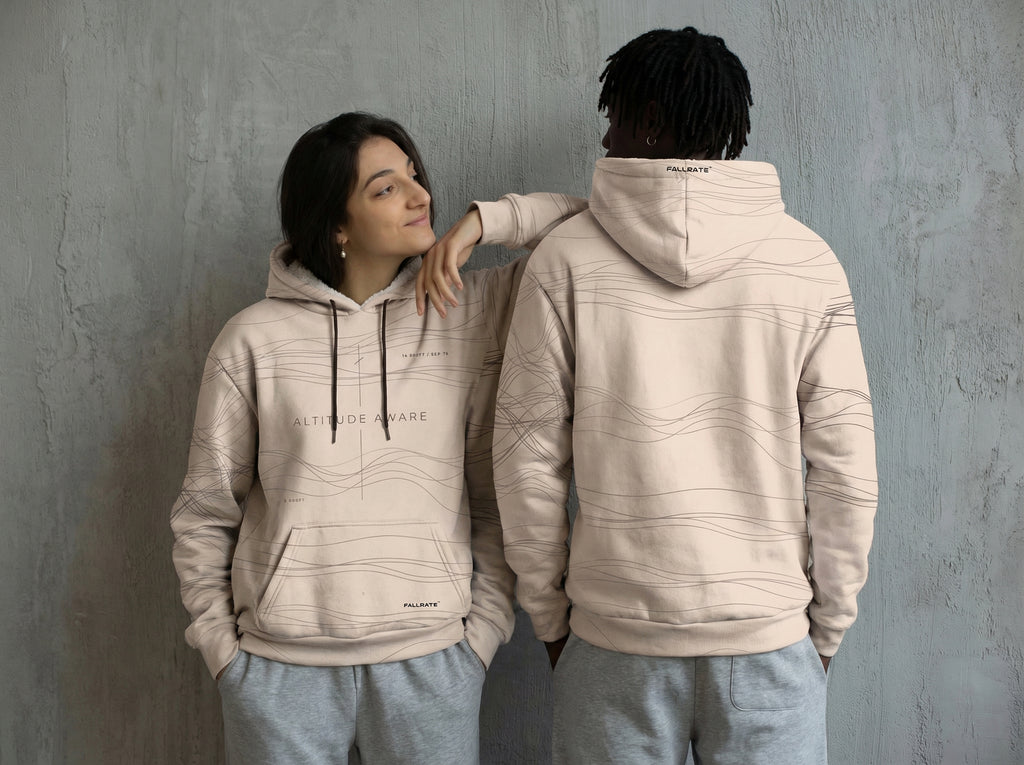 Two people wearing beige hoodies with a brand logo against a gray wall.