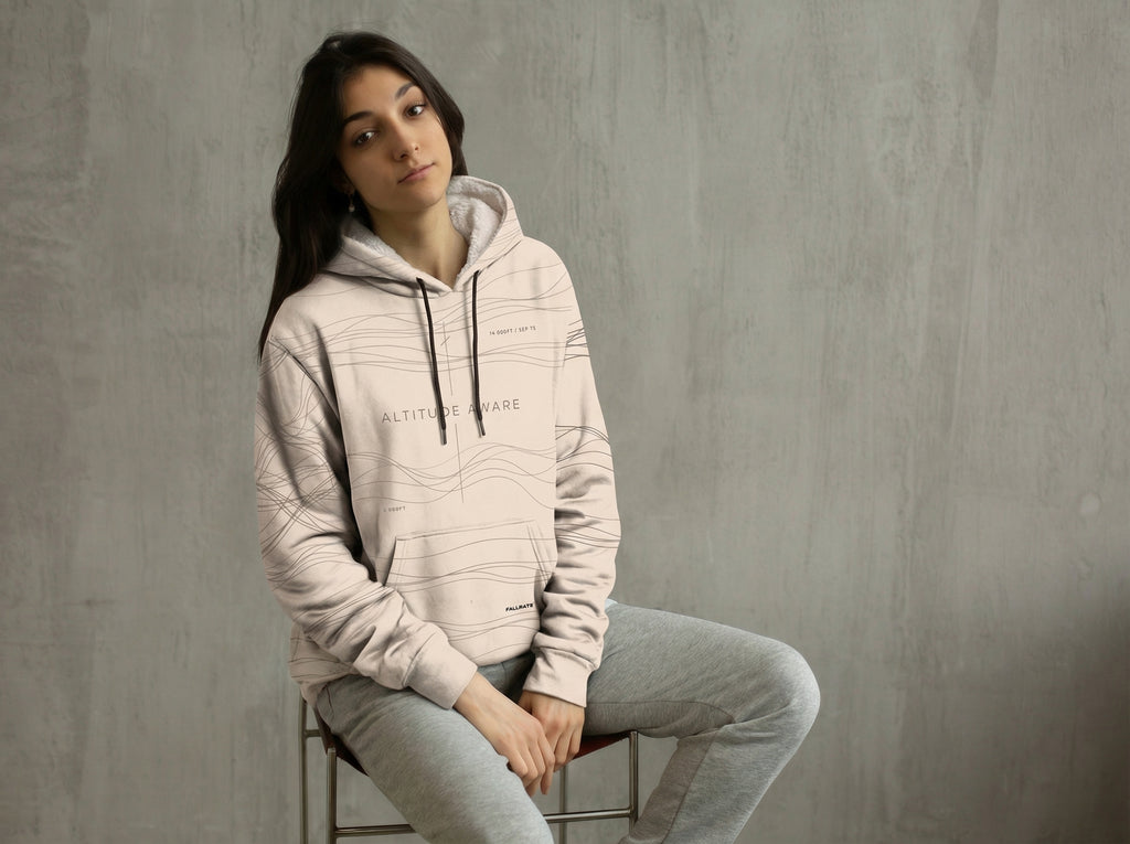 Woman wearing a light gray hoodie sitting on a stool against a plain wall.